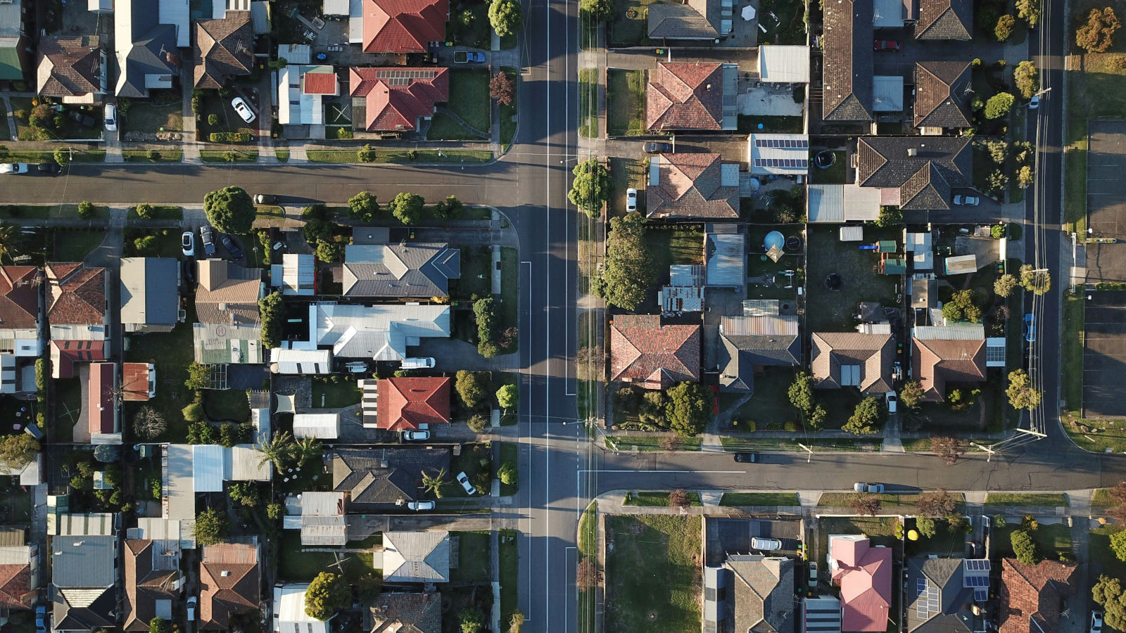 Aerial view of a suburban neighborhood with intersecting streets, houses with red and gray roofs, green lawns, and scattered trees.