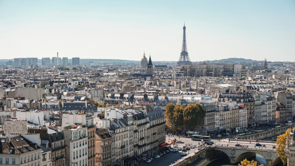 A panoramic view of Paris with the Eiffel Tower rising above the city’s classic rooftops and a bridge crossing the Seine River in the foreground.