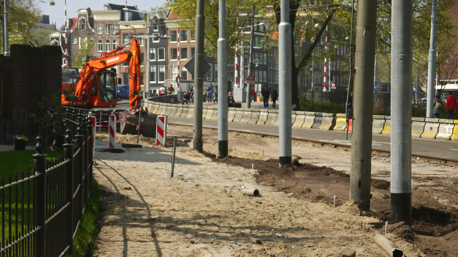 Construction site with an orange excavator, sandy ground, and poles, located next to a road with historic buildings in the background.