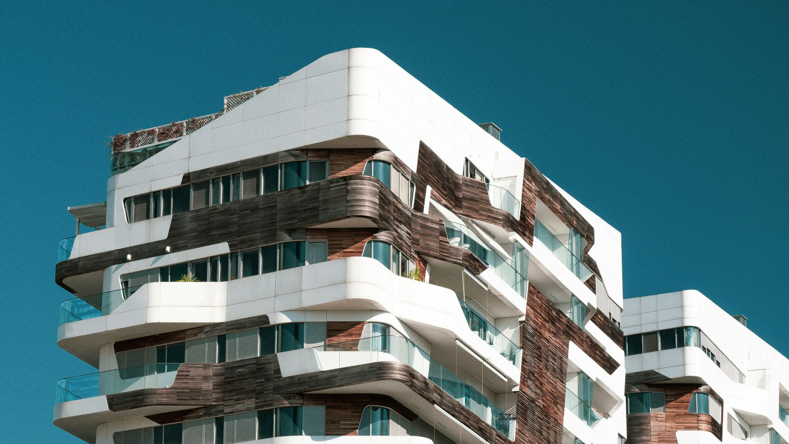 Modern apartment building with curved white and wooden facades, glass balconies, and large windows under a clear blue sky.